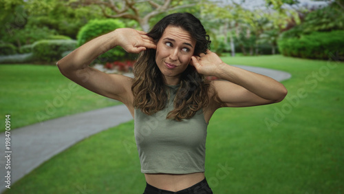 Bilde på lerret Young hispanic woman with bare stomach covers ears with hands in green park pathway under bright sunlight; irritation