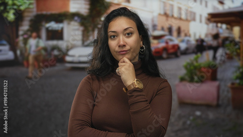 Foto Woman with hand on chin and hoop earring on a cobblestone street beside potted planters and parked cars wearing brown turtleneck; thoughtful confidence