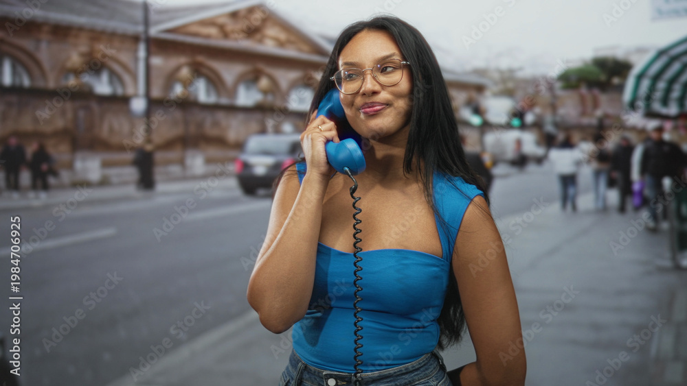Fototapeta premium Woman holding a blue corded telephone to her ear on a busy street near a historic building; contentment.