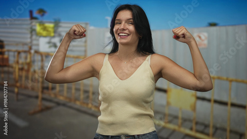 Принти на полотні Young hispanic woman flexes biceps while smiling at building under blue sky; empowerment confidence