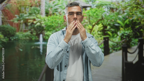 Foto Man covering mouth with hands in garden by pond and fountain, wearing glasses and denim jacket over white tshirt; surprise contemplation