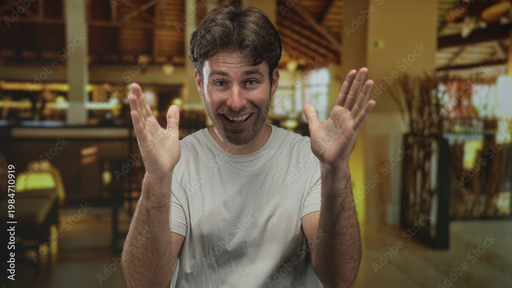 Naklejka premium Man smiling with hands raised and palms open in a hotel building lobby near reception desk and wooden beams; joyful humor candid.