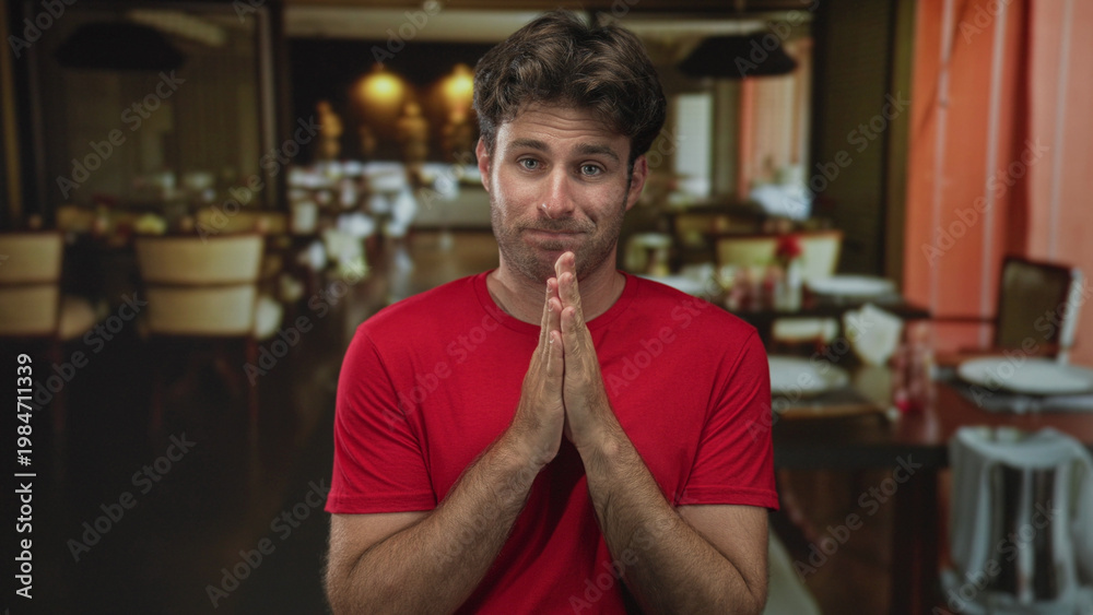 Fototapeta premium Man in red t shirt with hands clasped and grimace seated at a restaurant table inside a building; nervousness.