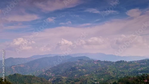 A wide panoramic view of terraced hills and scattered village homes under a vast sky filled with soft, drifting clouds in Mukteshwar, Uttarakhand, India.