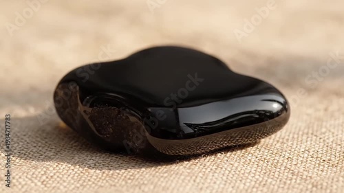 Smooth, glossy black stone with organic shape resting on a textured beige fabric surface, soft natural light creating gentle shadows