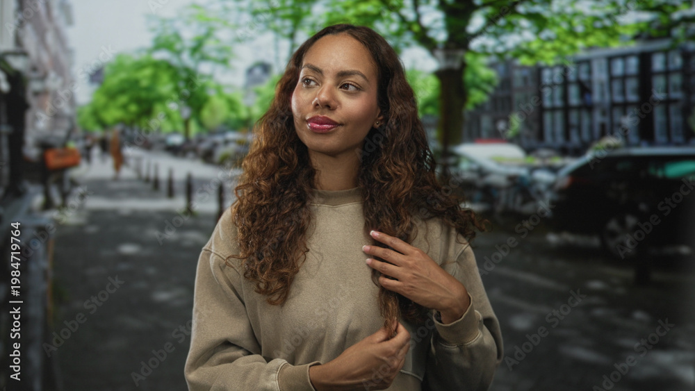 Fototapeta premium Young woman touches hair with both hands on a city street lined with trees and parked cars; confidence wellness.