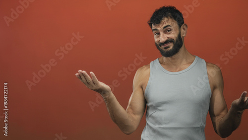 Photos Man showing palms up shrug with visible hand gesture, wearing a tanktop and beard against a red studio backdrop; bemusement acceptance