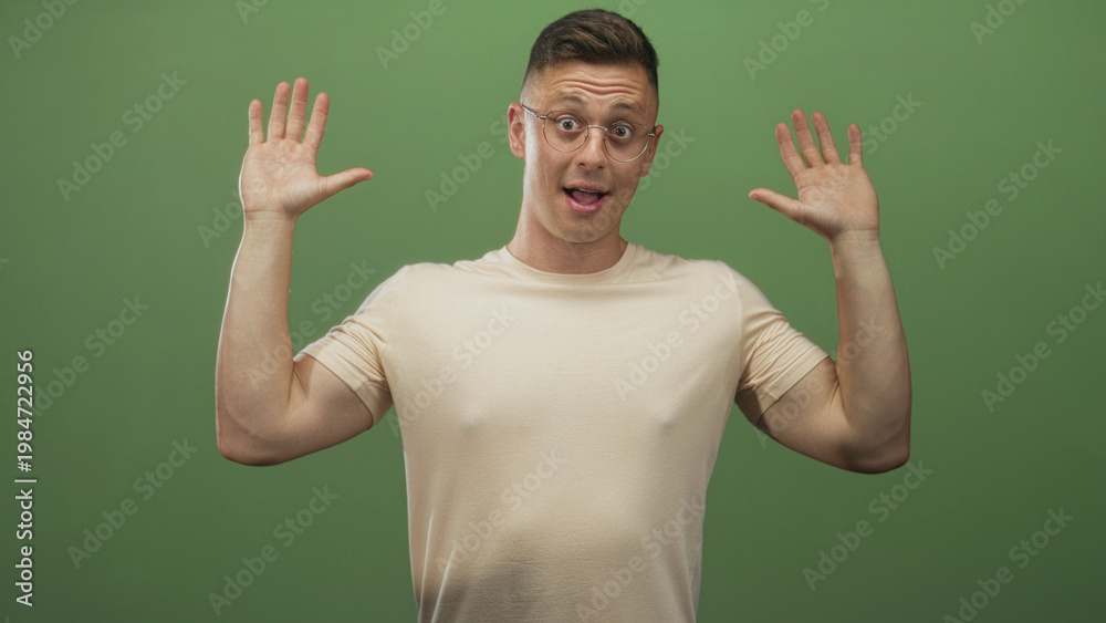 Fototapeta premium Man raising hands with palms up, smiling in beige tshirt and glasses against a green screen studio backdrop; confidence friendliness.