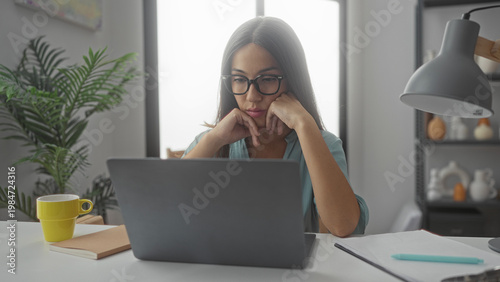 Wallpaper Mural Woman resting chin on hands while looking at laptop screen in home office with mug and plant on desk; contemplation focus. Torontodigital.ca