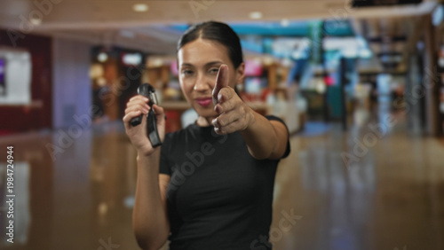 Εκτύπωση καμβά Young woman points finger like gun while holding a stun device at a mall kiosk, brunette in black shirt indoors center; self defense confidence