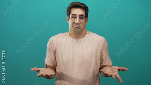 Obraz na płótnie Man shrugging with palms up and open hands in studio against teal backdrop, wearing glasses and pink tshirt; uncertainty