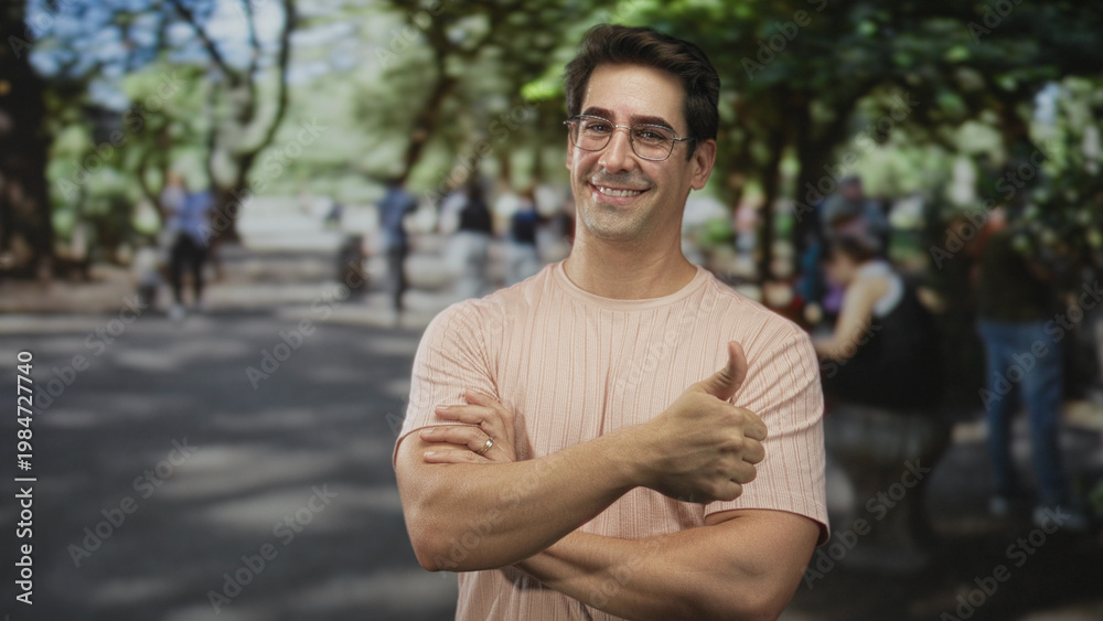 Fototapeta premium Man smiling with arms crossed and right hand giving thumbs up, wearing pink t shirt and glasses in street park with benches and trees visible; contentment.