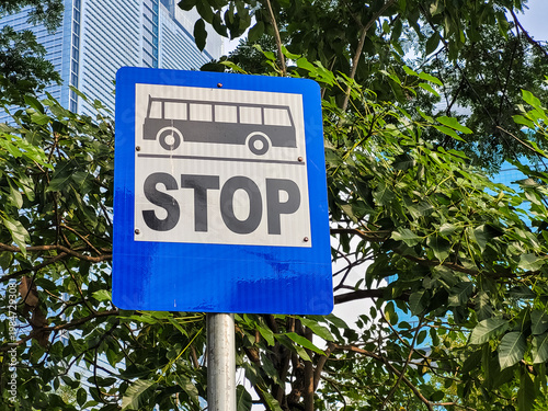 Bus stop sign with clear stop instruction placed along a city street, surrounded by green trees and modern buildings, representing urban transportation and public transit system.