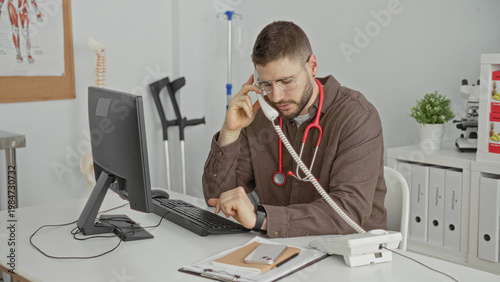 Wallpaper Mural Man doctor with stethoscope holds telephone to ear and types at computer in building; focused care coordination. Torontodigital.ca