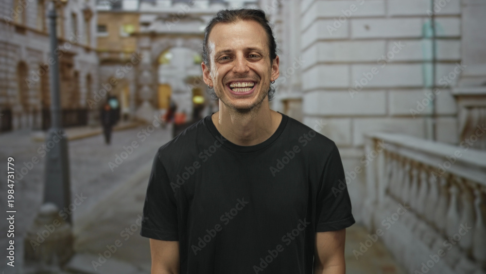 Obraz premium Man smiling with visible teeth and long hair tied back on city street beside stone balustrade and lamppost while wearing black t shirt; joy confidence.