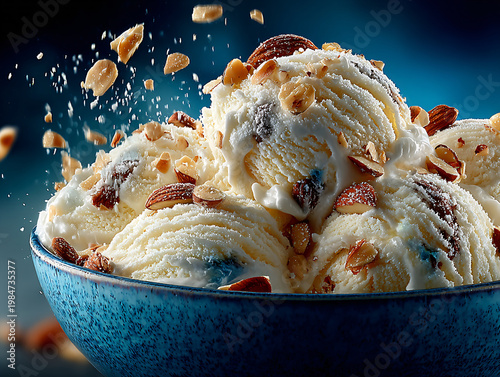 A generous mound of creamy white vanilla ice cream sits in a deep blue bowl, topped with crunchy almonds, drizzled with sweet sauce, and surrounded by flying nut pieces against a dark background.