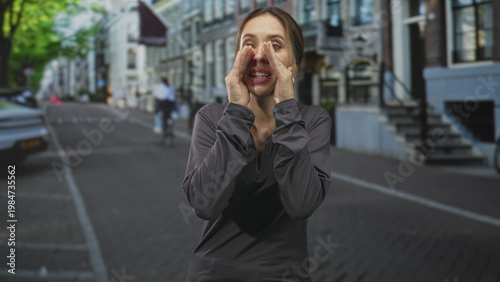 Obraz na plátně Woman cupping hands around mouth shouting to call attention on a city street with row houses and parked car; urgency warning