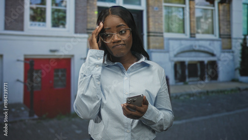 Bilde på lerret Woman holding smartphone with one hand and hand to forehead gesture while reading the screen in front of a building on a street; quiet concern