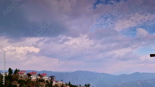 A dramatic sky filled with thick, layered clouds stretches over distant hills, with a cluster of red-roofed hillside homes perched on edge in Mukteshwar, Uttarakhand, India.