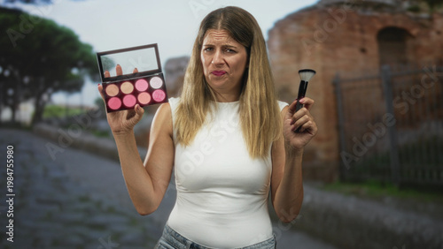 Photos Woman holds makeup palette and brush, frowning and raising eyebrows while wearing a white tanktop on a cobblestone street near ancient ruins; frustration doubt
