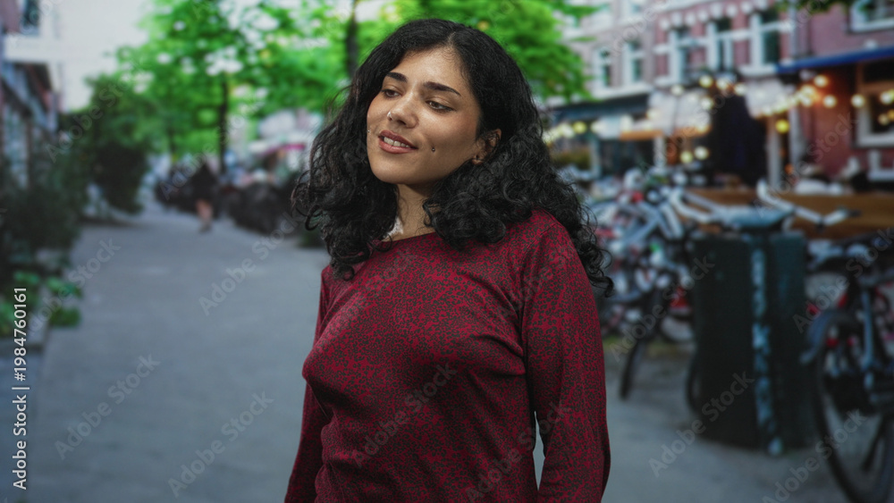 Fototapeta premium Young brunette woman smiling with face turned to side, wearing red sweater, standing near parked bicycles on a city street; serenity connection.