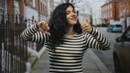 Fotografie Young woman on a street giving thumbs up with her right hand and thumbs down with her left, smiling in striped top; ambivalence contrast
