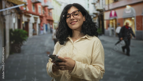 Foto Young brunette woman texting on smartphone with both hands visible while walking on a cobbled city street lined with shops; contentment connection