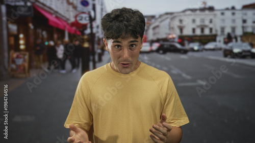 Φωτογραφία Young hispanic man clasping hands in prayer on a busy city street near storefronts and passing cars while looking upward; hopeful gratitude