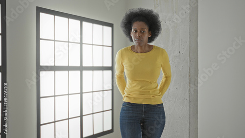 Obraz na plátně Woman in yellow sweater pulling out empty jeans pockets and shrugging beside large grid window in studio; financial worry