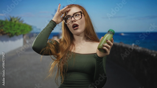 Canvas-taulu Redhead woman holding a green juice bottle with hand on forehead, shoulder exposed, making a reluctant sip gesture on a street; indifference