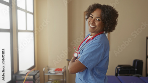 Wallpaper Mural Young african american doctor wearing blue scrubs with stethoscope smiles with crossed arms in modern clinic room; compassion. Torontodigital.ca