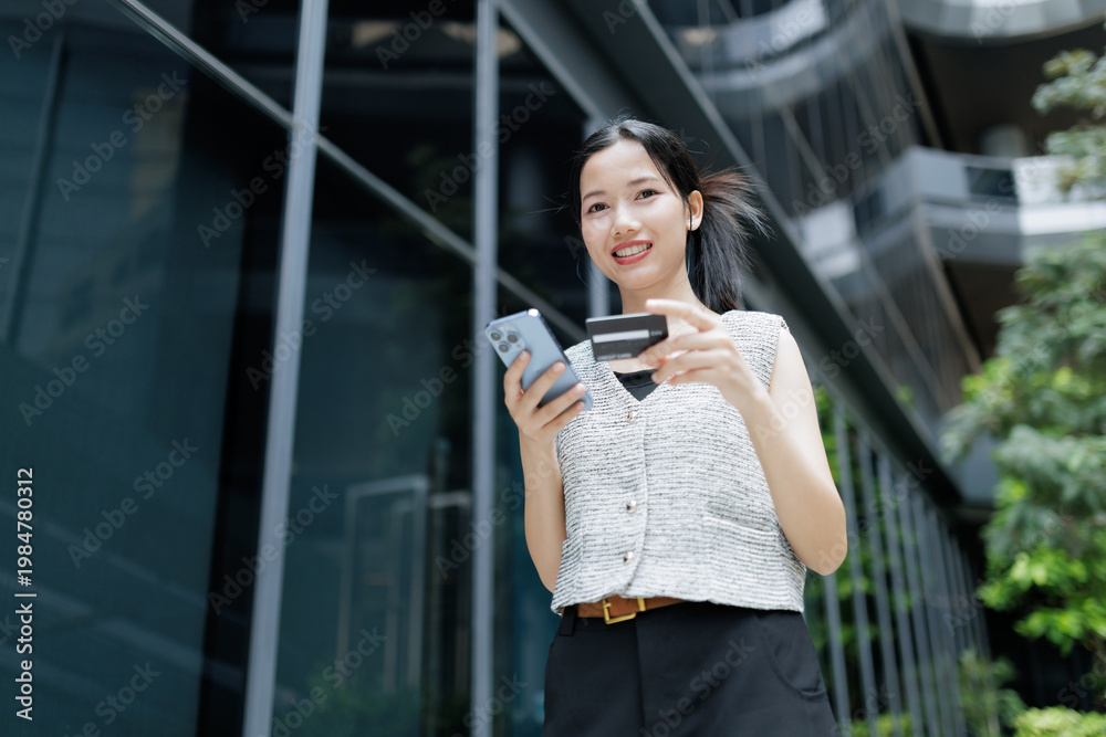 Fototapeta premium A young woman smiles while using her smartphone and holding a credit card outdoors near modern buildings.