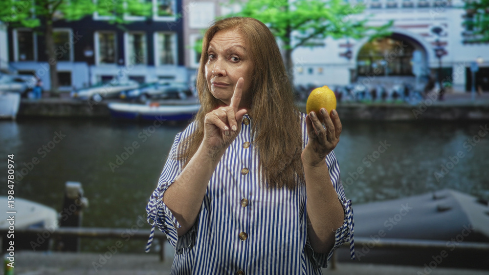 Fototapeta premium Woman holding lemon in left hand and points finger with right hand on a street by a canal; skepticism resilience.