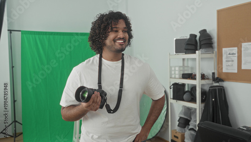 Man holding camera and smiling, hand on hip in studio with green screen and lighting gear; creative confidence.