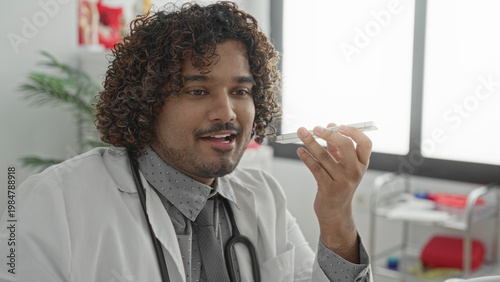 Bild auf Leinwand Young doctor man speaking into smartphone, holding phone to mouth, hand raised, in clinic building; compassionate care
