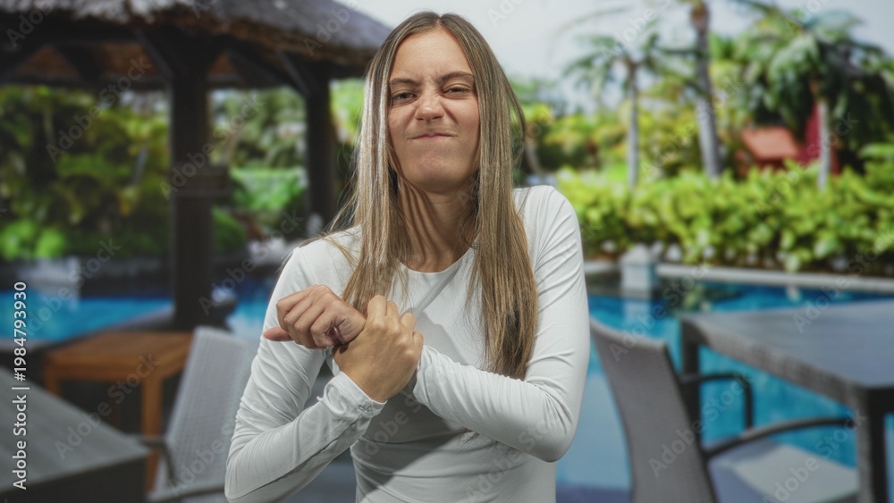 Fototapeta premium Woman clutching wrist and gripping her palm beside a hotel pool patio with lounge chairs and cabana; pain concern.