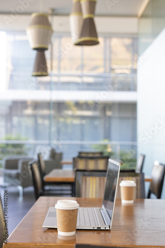 Open silver laptop sitting on wooden table with paper coffee cup near glass wall