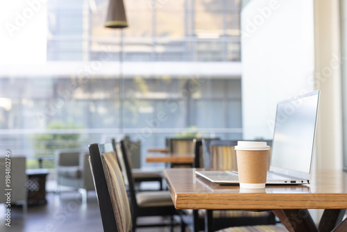 Paper coffee cup sitting on wooden table beside slim laptop at modern cafe, copy space