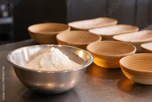 Stainless-steel mixing bowl holding mound of flour sitting left on countertop, bannetons visible