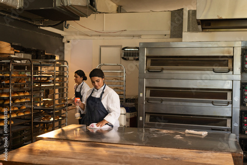 Female bakers cleaning prep table in chef coats using sprayer and organizing rack trays in bakery