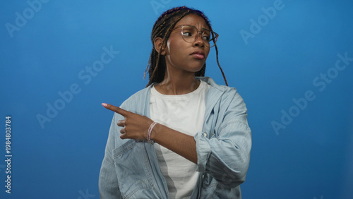 Tableau sur toile Woman pointing finger to the left in studio with blue backdrop, visible index finger and glasses, denim shirt and braided hair; disapproval