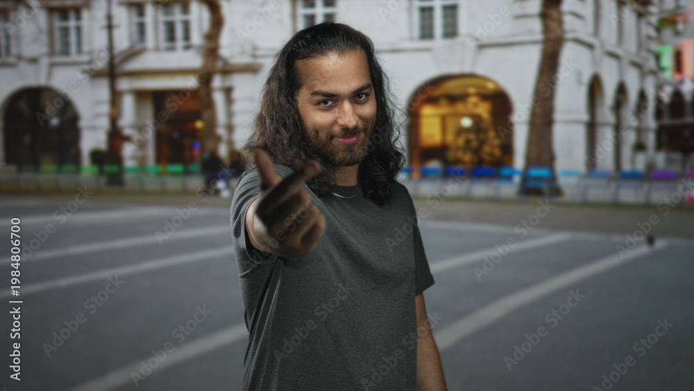 Naklejka premium Young man beckoning with extended hand and warm smile on a busy city street with historic building facades; friendly invitation.