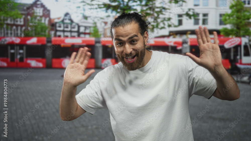 Naklejka premium Man with hands raised palms up wearing white t shirt on a cobbled city street in front of tram and shops, waving openly; friendly greeting happy.