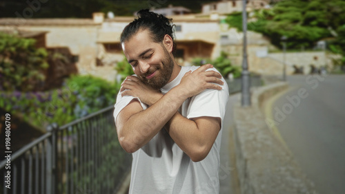 Cuadro en lienzo Young arab hispanic man hugging his arms across chest on a street near railing and buildings; self care serenity