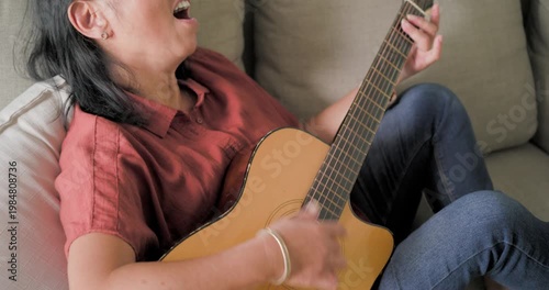 Woman is settling on light couch, starting to strum acoustic guitar and singing while relaxing