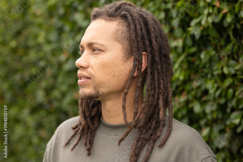 Mid-adult African American man standing and looking left by green hedge wearing olive sweatshirt