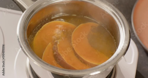 As white burner is heating, steel saucepan is simmering three orange squash slices softening edges