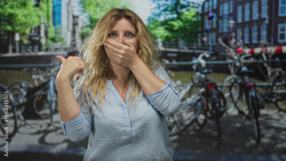 Fototapeta premium Woman with hand covering mouth and thumb pointing back on street by amsterdam canal with bicycles and brick buildings; surprise.