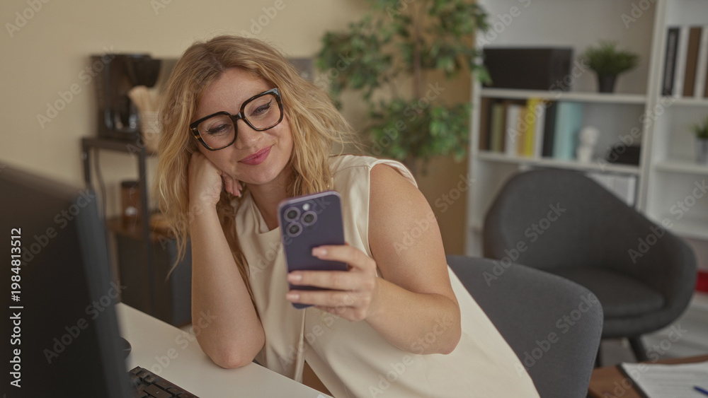 Fototapeta premium Woman wearing glasses holding smartphone, smiling while seated at a desk in an office interior beside a computer monitor; happiness work balance.