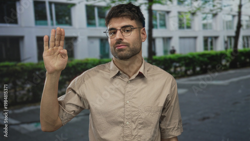 Foto Man in beige shirt and glasses raising hand in greeting on sunlit tree lined city street; friendly greeting